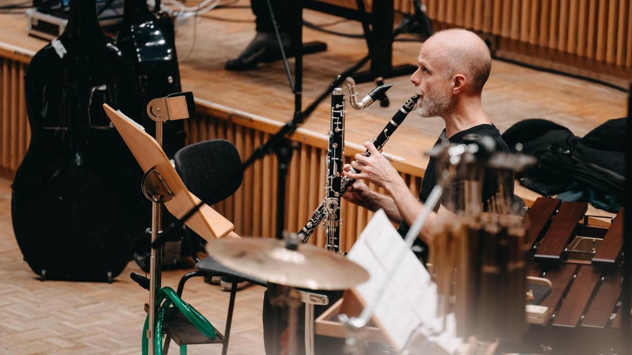 Ein Musiker mit einer Klarinette sitzt hochkonzentriert zwischen Pulten und auf einer Bühne und spielt. Ein Musiker mit einer Klarinette sitzt hochkonzentriert zwischen Pulten und auf einer Bühne und spielt.