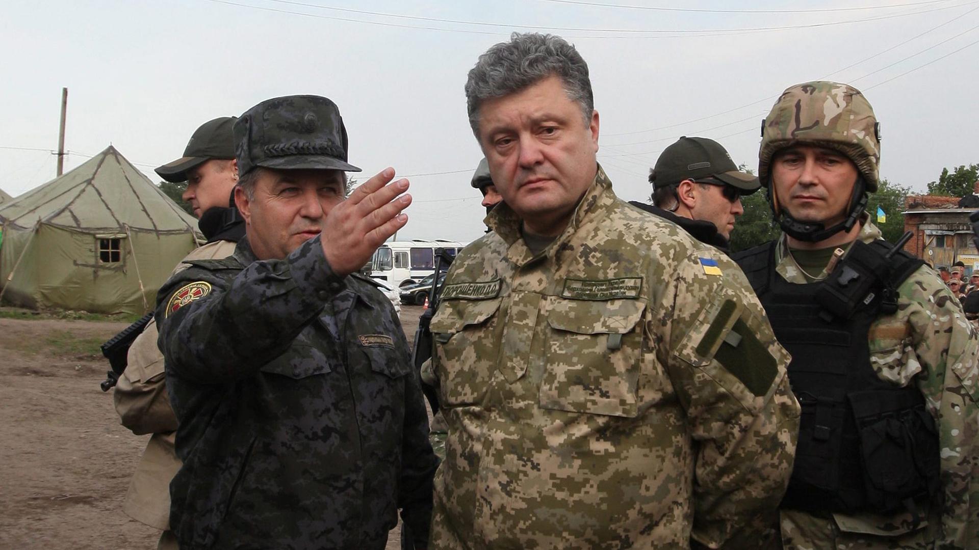 Der ukrainische Präsident Petro Poroschenko steht im Feldanzug gekleidet in einem Militärlager. Neben ihm ein Soldat, der mit ausgestrecktem Arm auf etwas außerhalb des Bildes deutet. Der ukrainische Präsident Petro Poroschenko steht im Feldanzug gekleidet in einem Militärlager. Neben ihm ein Soldat, der mit ausgestrecktem Arm auf etwas außerhalb des Bildes deutet.