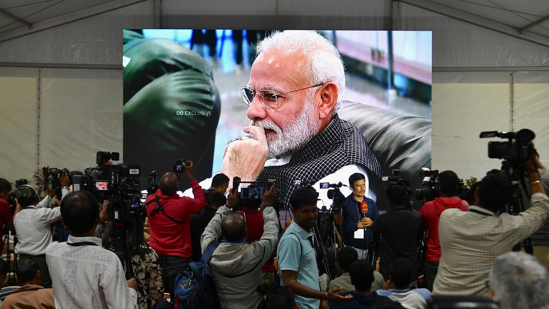 Indiens Premierminister Modi, zu sehen auf einer Leinwand, die in einem Zelt aufgestellt wurde. Davor Medienvertreter, welche die Landung der Mondsonde "Vikram" verfolgten.