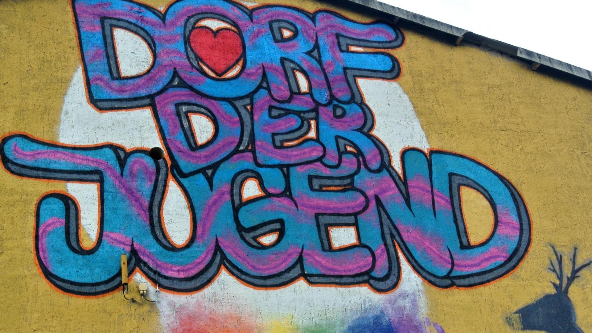 Schriftzug "Dorf der Jugend" in Blau und Pink an eine Mauer gesprayt, im "o" ein rotes Herz, im Hintergrund eine Wolke, aus der ein Regenbogen kommt, sowie ein Hirschkopf Schriftzug "Dorf der Jugend" in Blau und Pink an eine Mauer gesprayt, im "o" ein rotes Herz, im Hintergrund eine Wolke, aus der ein Regenbogen kommt, sowie ein Hirschkopf