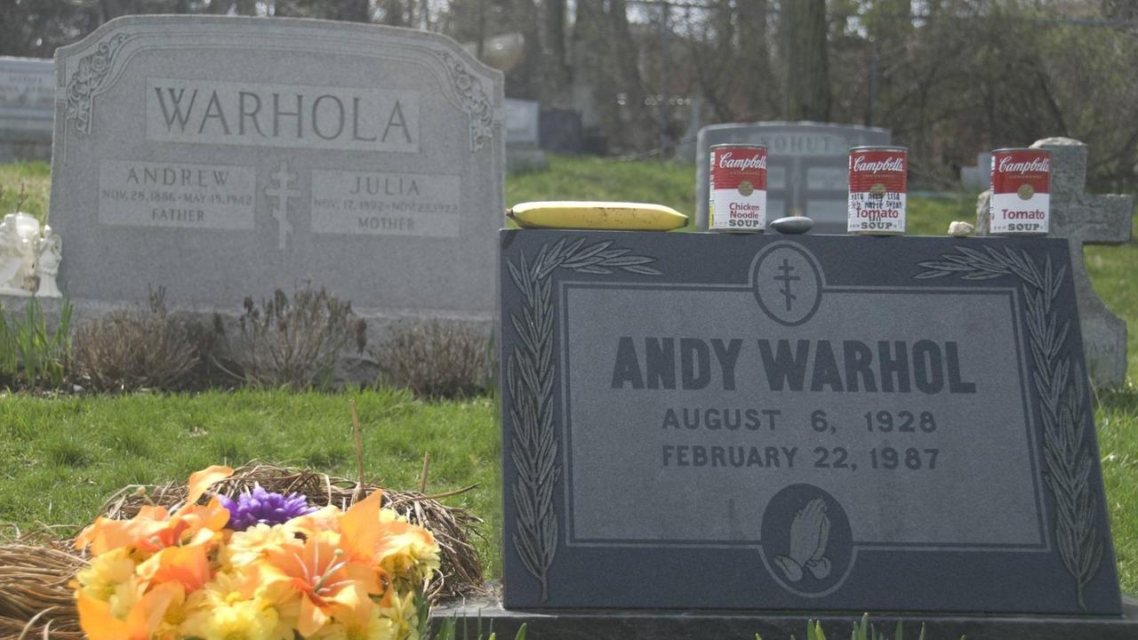 Das Bild zeigt Warhols Grabstein, auf den neben einer Banane, mehrere Dosen Campbells Tomatensuppe gestellt wurden. Im Hintergrund ist das Grab bzw. der Grabstein seiner Eltern zu sehen.
