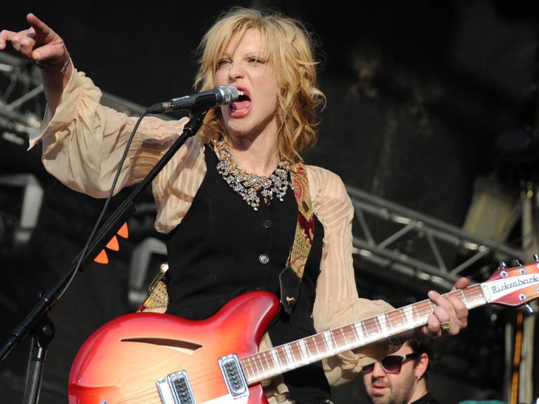 Eine grimmige Courtney Love bei einem Konzert in Moskau: Mit ironsich edler Weste, weiten Ärmeln, Perlenschmuck und einer knallroten E-Gitarre.