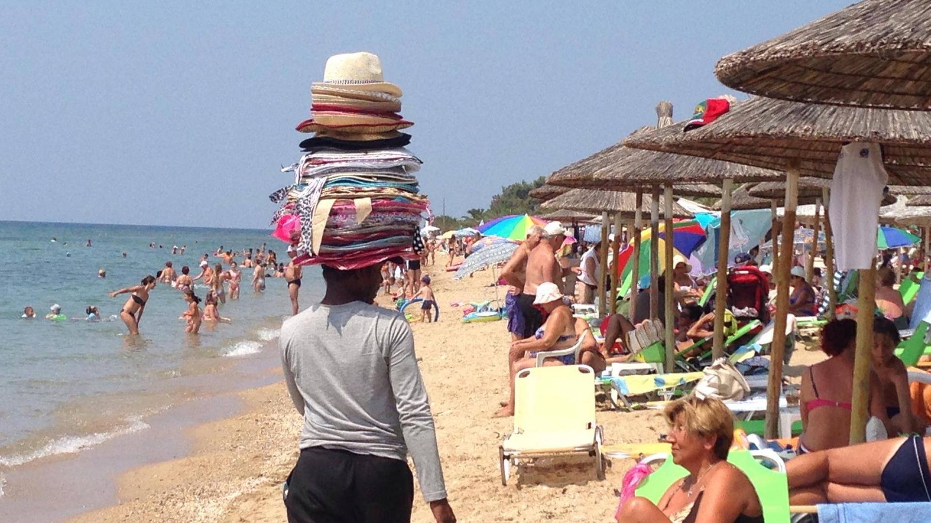 Ein Mann von hinten, mit vielen Hüten gestapelt auf dem Kopf, schlendert am Strand entlang. Er ist ein Verkäufer, ein fliegender Händler, der versucht, das Interesse der unter Schirmen sitzenden Strandbesucher zu wecken. Ein Mann von hinten, mit vielen Hüten gestapelt auf dem Kopf, schlendert am Strand entlang. Er ist ein Verkäufer, ein fliegender Händler, der versucht, das Interesse der unter Schirmen sitzenden Strandbesucher zu wecken.