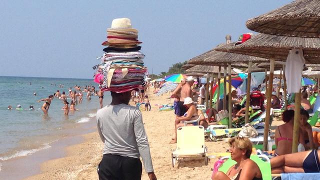 Ein Mann von hinten, mit vielen Hüten gestapelt auf dem Kopf, schlendert am Strand entlang. Er ist ein Verkäufer, ein fliegender Händler, der versucht, das Interesse der unter Schirmen sitzenden Strandbesucher zu wecken.