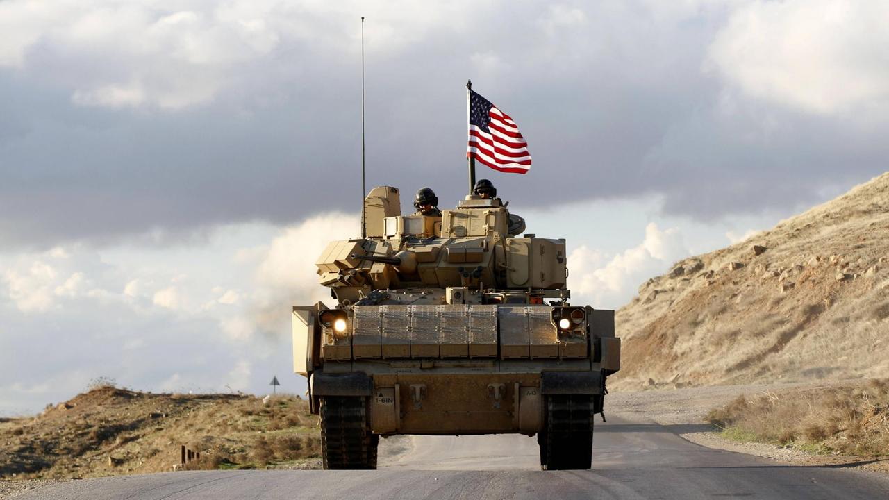 Ein Patrouillenfahrzeug der Joint Task Force CJTF-OIR mit US-Soldaten fährt auf einer Wüstenstraße. Oben auf dem Wagen weht eine US-Flagge.