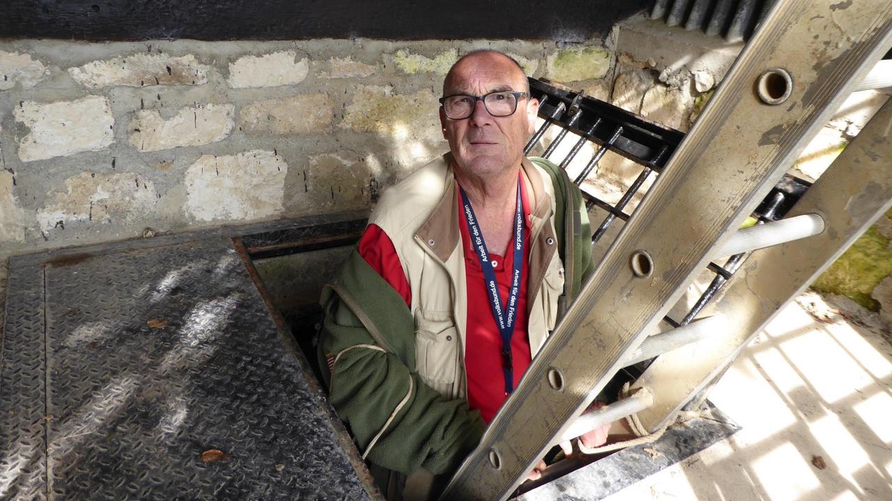 Auf einer Leiter, die in ein Kellergewölbe führt, steht ein älterer Mann mit rotem Shirt und Brille und schaut nach oben Richtung Kamera. Auf einer Leiter, die in ein Kellergewölbe führt, steht ein älterer Mann mit rotem Shirt und Brille und schaut nach oben Richtung Kamera.