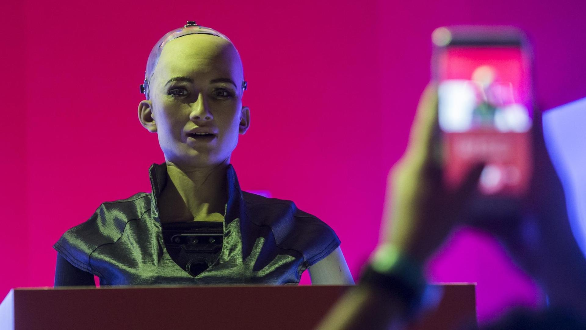 "Sophia the Robot" steht hinter einem Podium, mit einer pinken Fläche im Hintergrund und wird mit einem Smarphone fotografiert. "Sophia the Robot" steht hinter einem Podium, mit einer pinken Fläche im Hintergrund und wird mit einem Smarphone fotografiert.