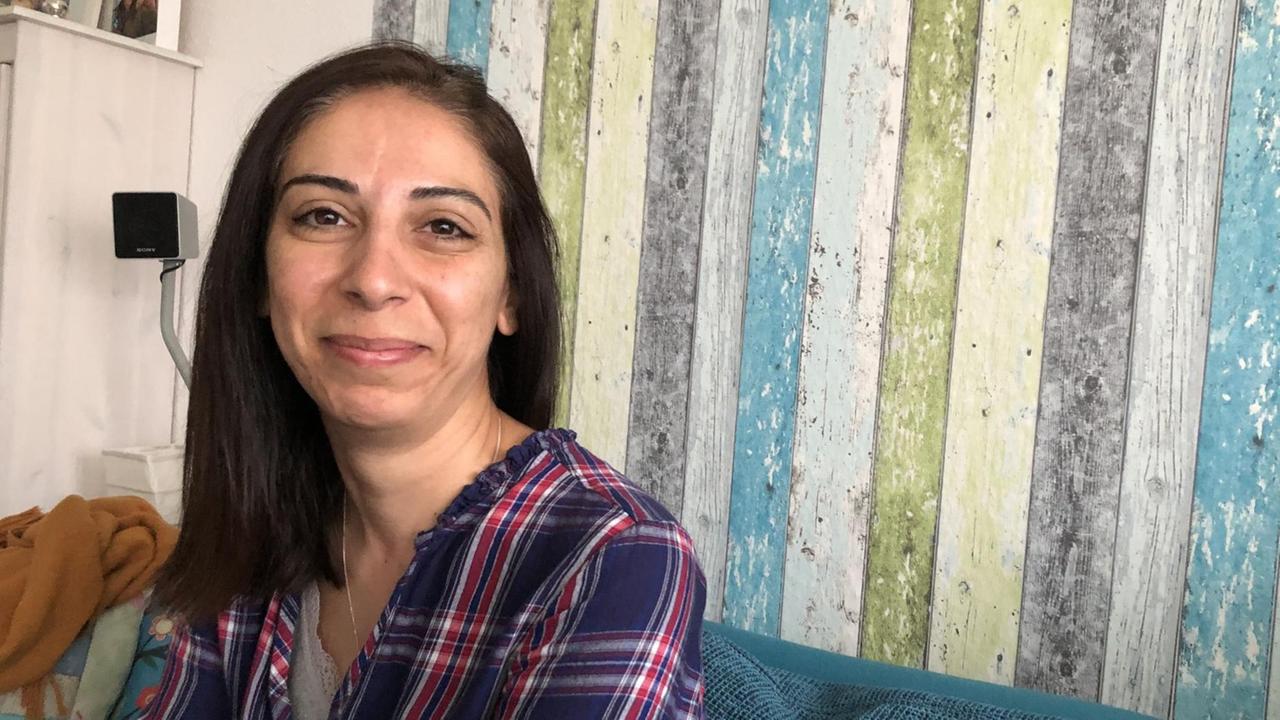Eine Frau mit langen glatten dunklen Haaren und kariertem Hemd sitzt auf einem Sofa und lächelt in die Kamera. Eine Frau mit langen glatten dunklen Haaren und kariertem Hemd sitzt auf einem Sofa und lächelt in die Kamera.