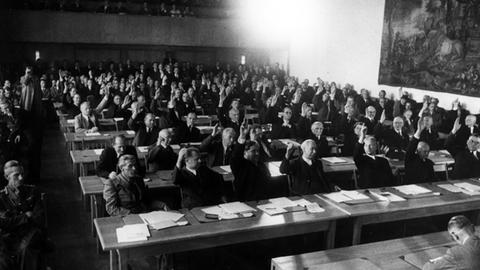 Ein Schwarz-Weiß-Foto zeigt die Annahme des Grundgesetzes 1949. Das Plenum des Parlamentarischen Rates nimmt in 3. Lesung am 8. Mai 1949 den revidierten Grundgesetzentwurf mit 53 gegen 12 Stimmen an. - 1.Reihe v.l.: Max Reimann, Walter Menzel, Carlo Schmid, Theodor Heuss.