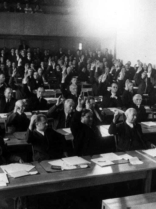 Ein Schwarz-Weiß-Foto zeigt die Annahme des Grundgesetzes 1949. Das Plenum des Parlamentarischen Rates nimmt in 3. Lesung am 8. Mai 1949 den revidierten Grundgesetzentwurf mit 53 gegen 12 Stimmen an. - 1.Reihe v.l.: Max Reimann, Walter Menzel, Carlo Schmid, Theodor Heuss.