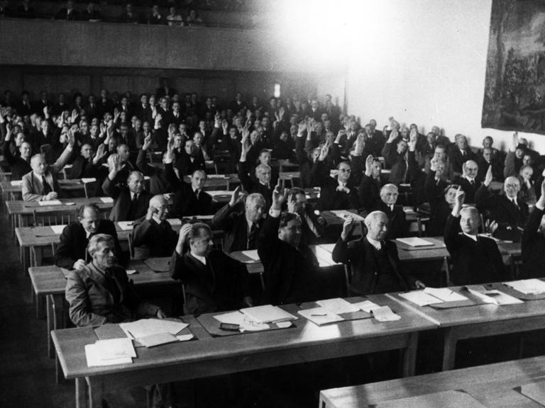 Ein Schwarz-Weiß-Foto zeigt die Annahme des Grundgesetzes 1949. Das Plenum des Parlamentarischen Rates nimmt in 3. Lesung am 8. Mai 1949 den revidierten Grundgesetzentwurf mit 53 gegen 12 Stimmen an. - 1.Reihe v.l.: Max Reimann, Walter Menzel, Carlo Schmid, Theodor Heuss.