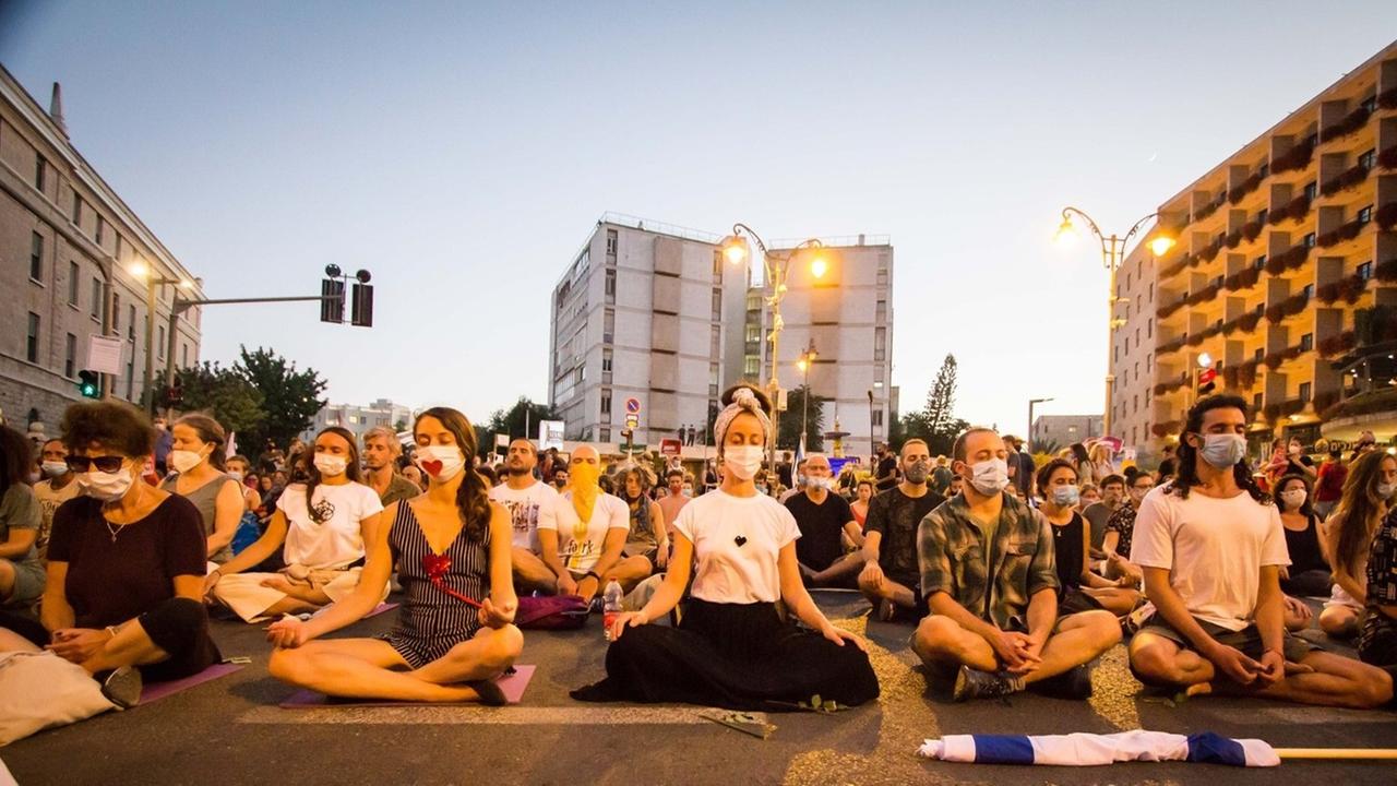 Mit Mundschutz sitzen Menschen im Schneidersitz auf einer Straßenkreuzung und praktizieren Yoga. Mit Mundschutz sitzen Menschen im Schneidersitz auf einer Straßenkreuzung und praktizieren Yoga.