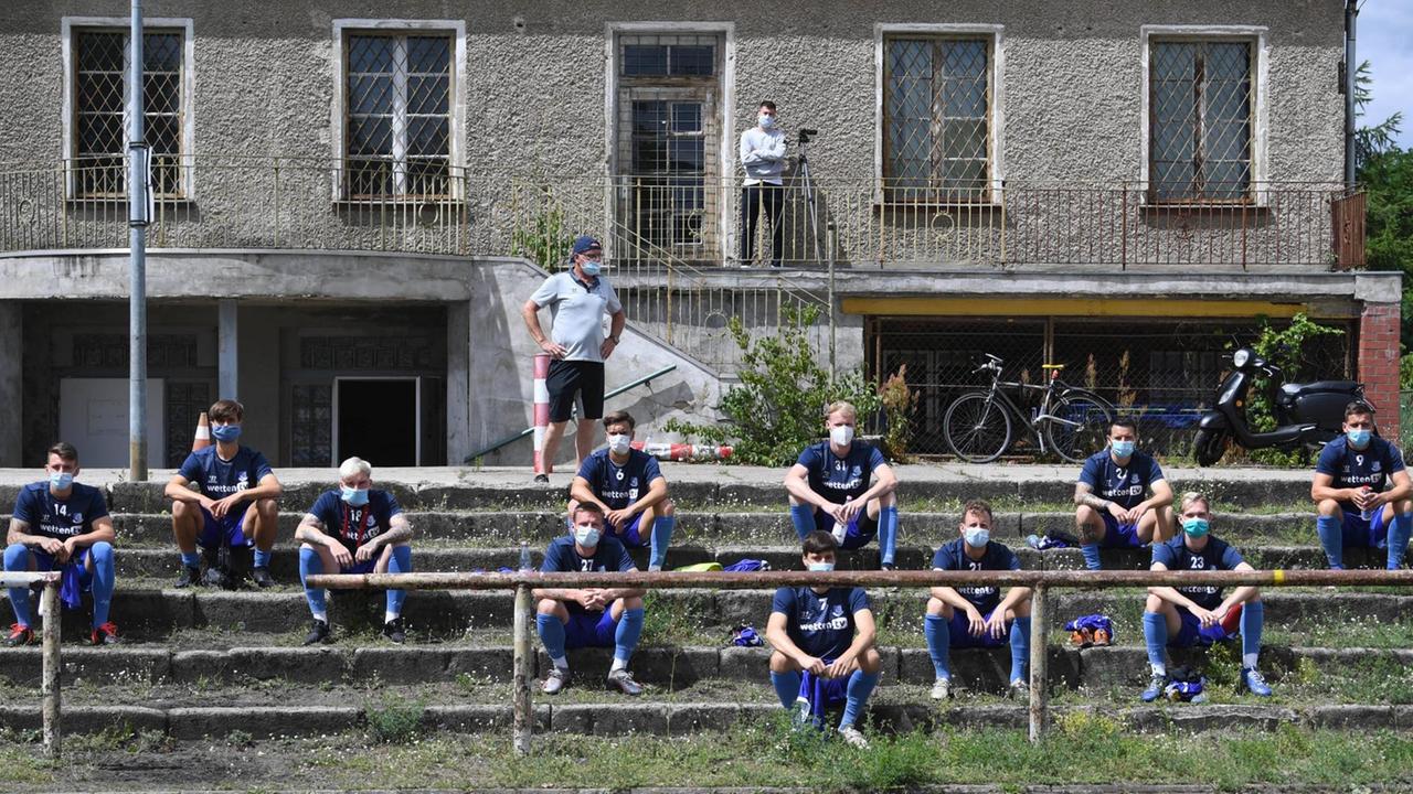 Fußballspieler sitzen mit Abstand auf einer Betontribüne und tragen grüne Gesichtsmasken. Fußballspieler sitzen mit Abstand auf einer Betontribüne und tragen grüne Gesichtsmasken.