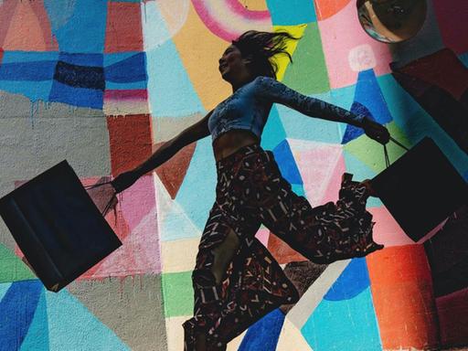 Eine Frau springt mit ihren Einkaufstüten tänzerisch vor einer bunten Fassade.