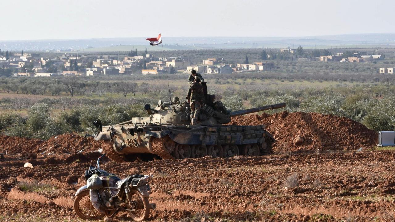 Der Panzer und ein Motorrad stehen auf einer schlammigen braunen Fläche. Auf dem Panzer stehen zwei Soldaten. Im Hintergrund das Panorama der Stadt.