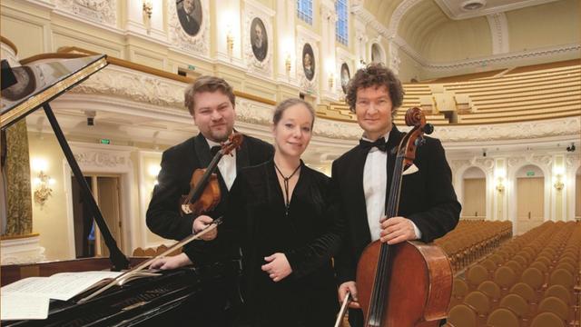 Drei Personen in schwarzer Kleidung sthen zusammen in einem Konzertsaal neben einem Flügel. Die beiden Männer halten je ein Instrument in den HÄnden, eine Violine und ein Cello.