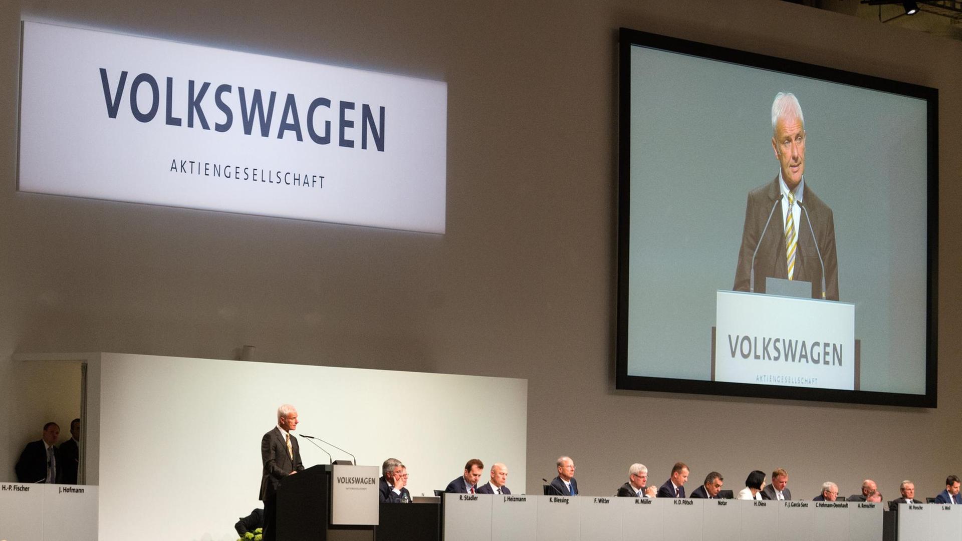 Man sieht Müller klein am Rednerpult und groß auf der Videowand dahinter. Neben ihm sitzen die Volrstände und Aufsichtsräte. Man sieht Müller klein am Rednerpult und groß auf der Videowand dahinter. Neben ihm sitzen die Volrstände und Aufsichtsräte.