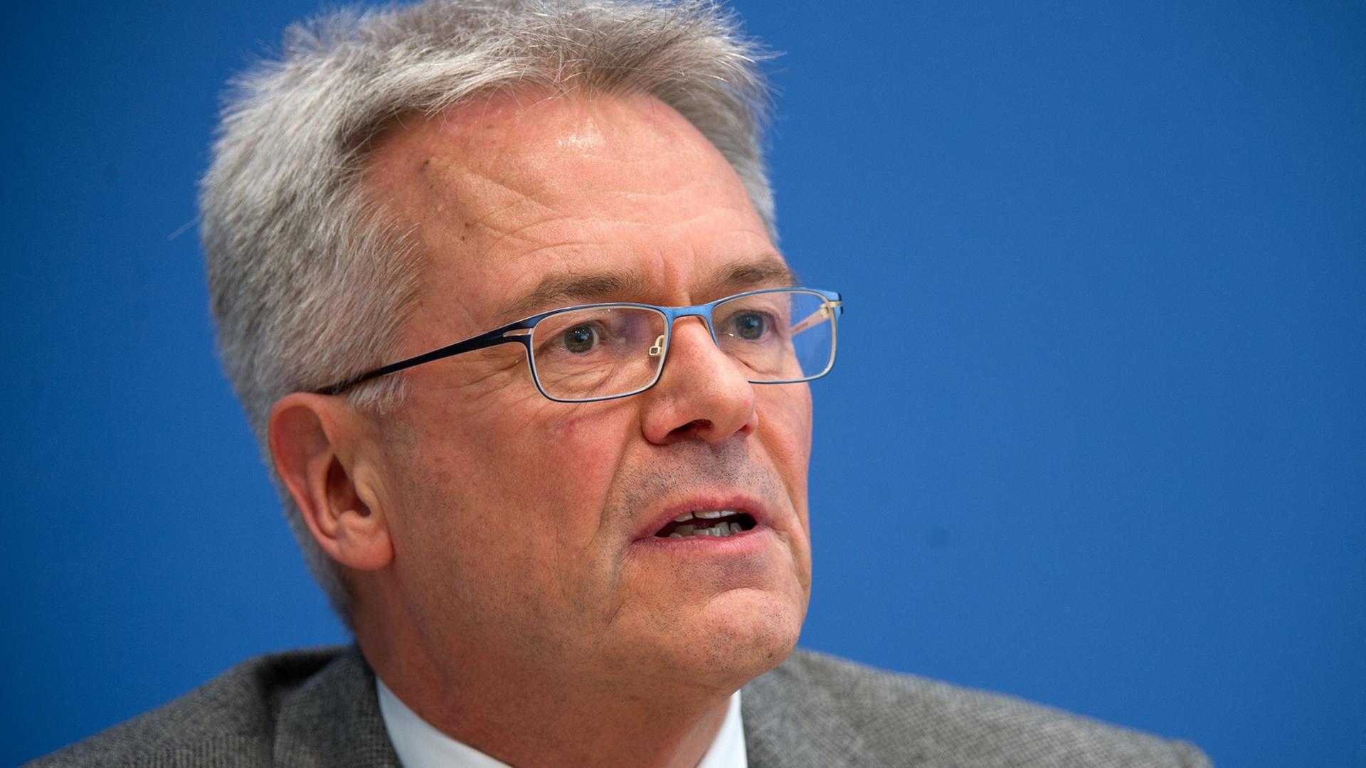 Mann mit grauen Harren, Brille und Anzug und Krawatte sitzt vor einer blauen Wand und spricht, Ausschnitt bis zu den Schultern Mann mit grauen Harren, Brille und Anzug und Krawatte sitzt vor einer blauen Wand und spricht, Ausschnitt bis zu den Schultern
