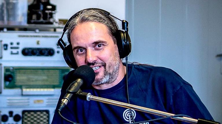 Ein Mann mit Kopfhörern sitzt in einem Raum voller technischer Anmutung und gibt ein Interview. Ein Mann mit Kopfhörern sitzt in einem Raum voller technischer Anmutung und gibt ein Interview.