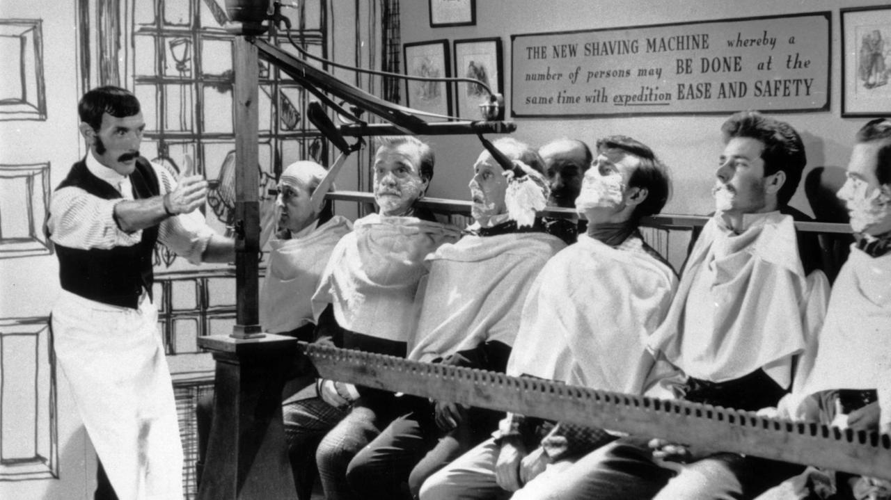 Der englische Komiker Eric Sykes (1923 - 2012, links) lässt in einer nicht ausgestrahlten Pilotsendung für eine geplante Fernsehserie namens "Brainwaves" am 28. Oktober 1960 die "Massenrasiermaschine" wieder aufleben, eine Erfindung aus dem 19. Jahrhundert, mit der ein Dutzend Männer gleichzeitig rasiert werden können. 
