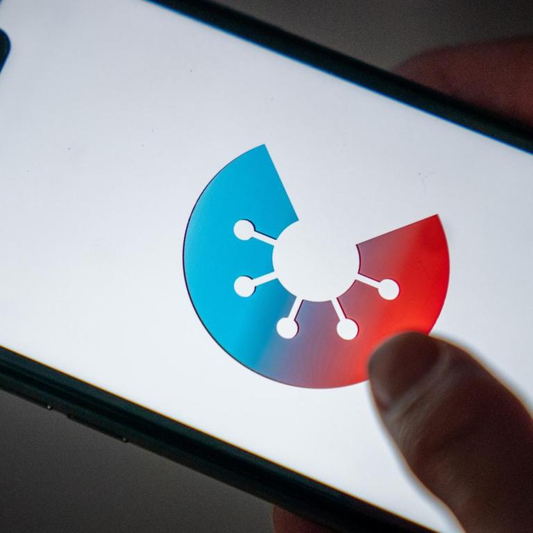 Zu sehen sind Hände, die ein Smartphone halten. Auf dem Display wird das rot-blaue Logo der Corona-Warn-App des Bundes angezeigt.