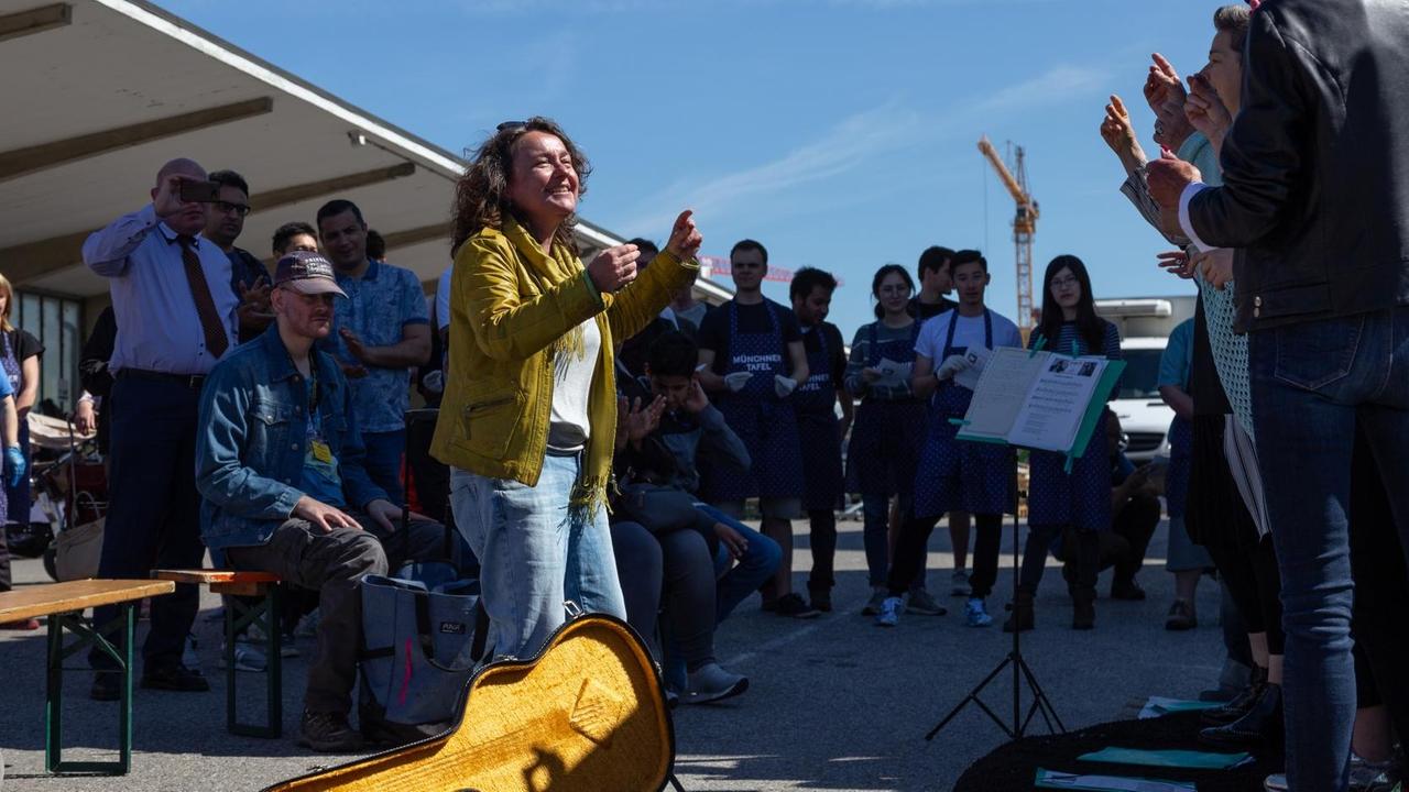 Eine Frau in gelber Jacke steht vor einem gelb ausgeschlagenen Gitarrenkasten und animiert um sie herumstehende Sängerinnen im Rhythmus zu bleiben. Eine Frau in gelber Jacke steht vor einem gelb ausgeschlagenen Gitarrenkasten und animiert um sie herumstehende Sängerinnen im Rhythmus zu bleiben.
