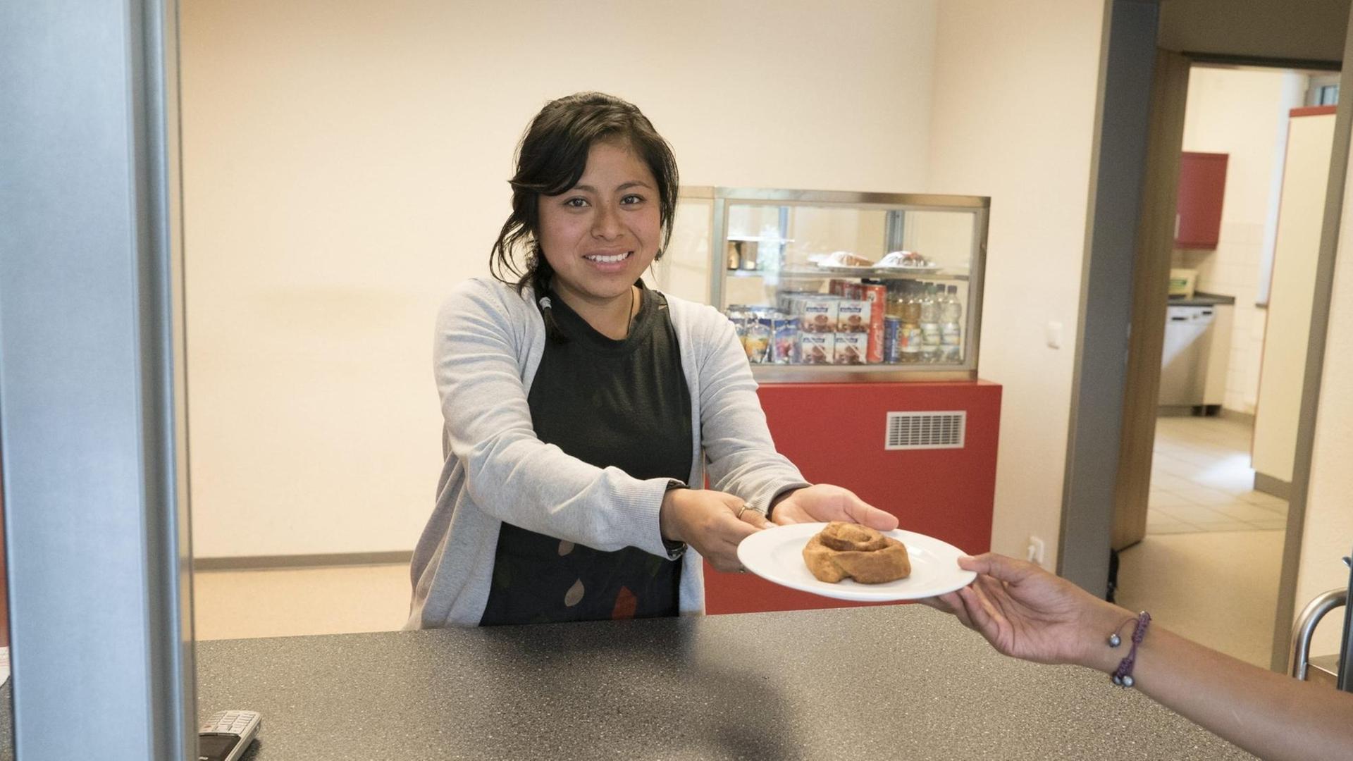 Eine junge Frau steht hinter einer Theke und reicht jemandem einen Teller mit einem Kuchenteilchen. Eine junge Frau steht hinter einer Theke und reicht jemandem einen Teller mit einem Kuchenteilchen.