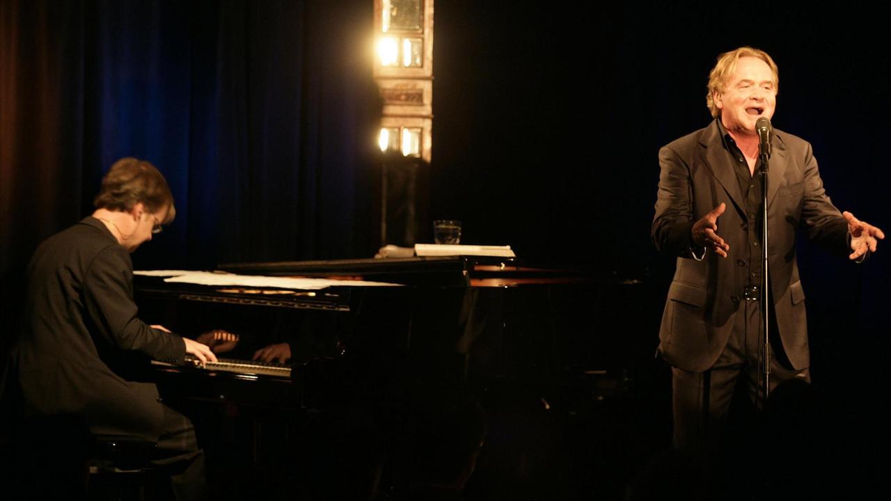 Klaus Hoffmann steht rechts auf einer dunklen Bühne, Hawo Bleich sitzt links am Piano.