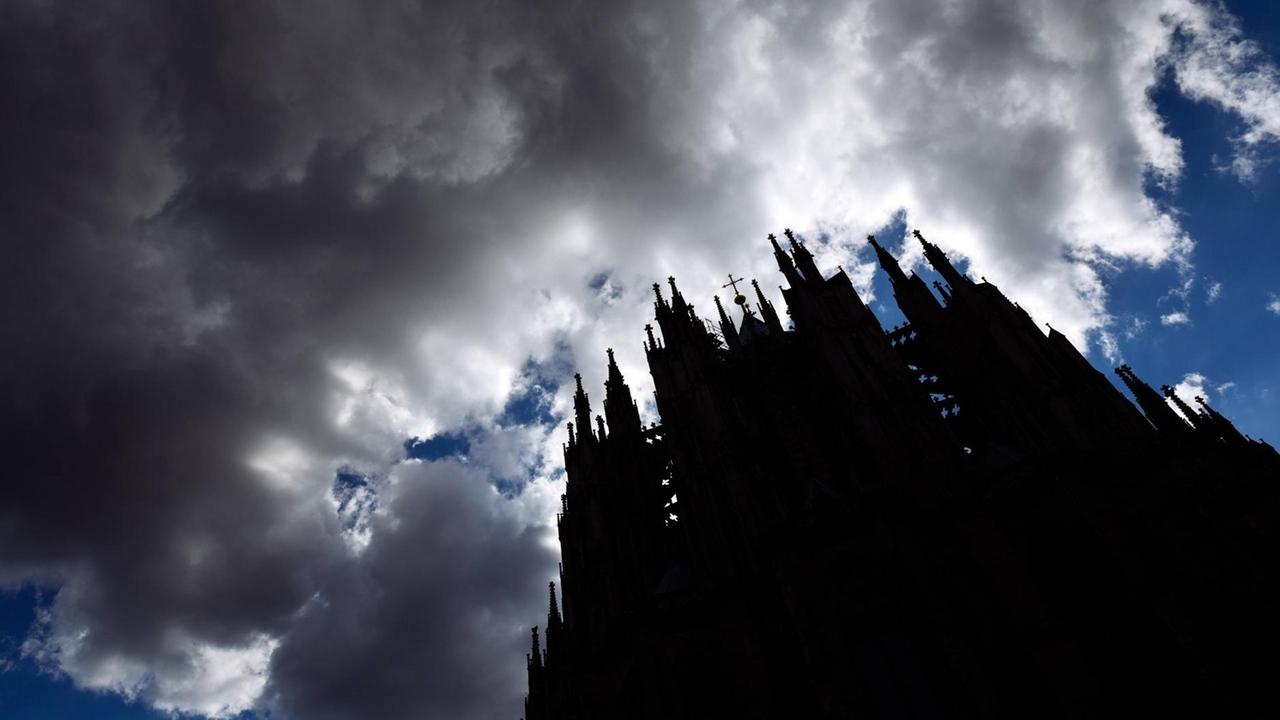Sturmwolken ziehen auf über dem Kölner Dom. Das Gebäude ist aus der Froschperspektive nur als Schattenriss erkennbar, darüber brauen sich dunkle Wolken zusammen.