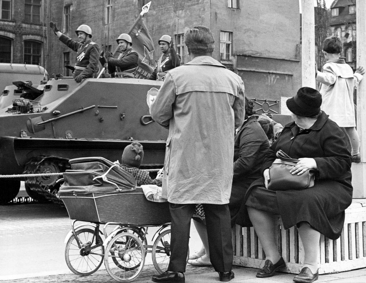 Das Schwarz-Weiß-Foto zeigt Zzuschauer einer Parade, im Hintergrund Soldaten in Uniform auf einem vorbeifahrenden Panzer. Das Schwarz-Weiß-Foto zeigt Zzuschauer einer Parade, im Hintergrund Soldaten in Uniform auf einem vorbeifahrenden Panzer.