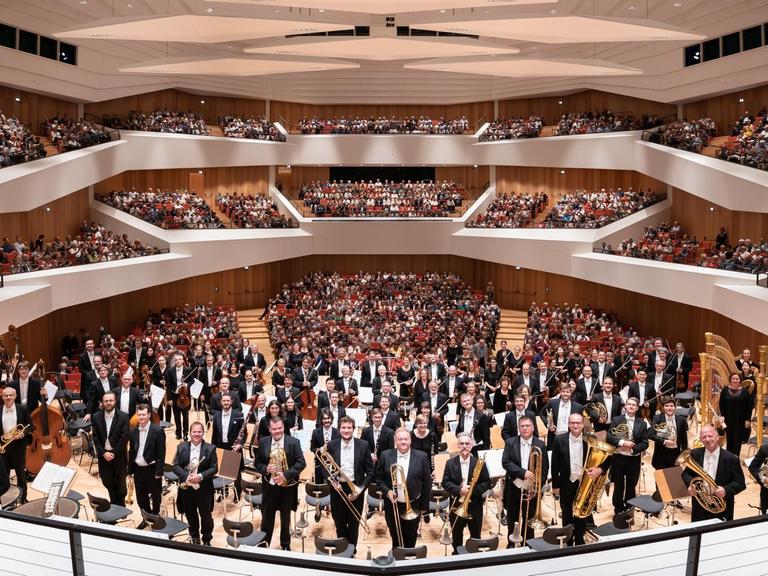 Ein Orchester steht vor einem voll besetzten Konzerthaus.