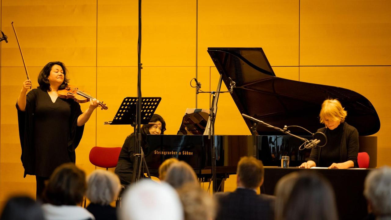 Eine Geigerin, eine Pianistin und eine Schauspielerin performen auf einer Bühne vor Publikum. Im Hintergrund ist eine gelbe Wand zu sehen. Eine Geigerin, eine Pianistin und eine Schauspielerin performen auf einer Bühne vor Publikum. Im Hintergrund ist eine gelbe Wand zu sehen.