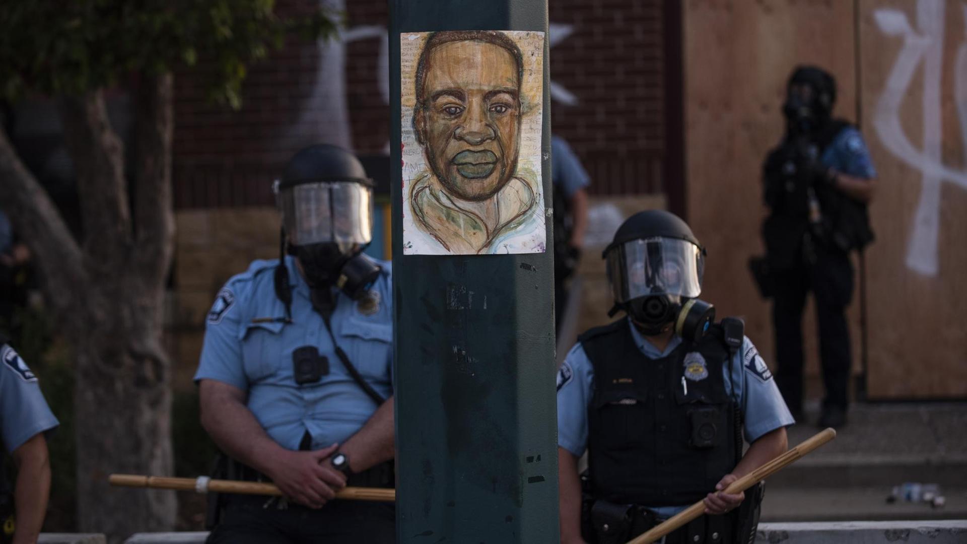 Ein Porträt von George Floyd hängt an einem Straßenlaternenmast, während Polizeibeamte bei einer Gegenüberstellung mit einer Gruppe von Demonstranten in Minneapolis, Minnesota, Wache stehen.