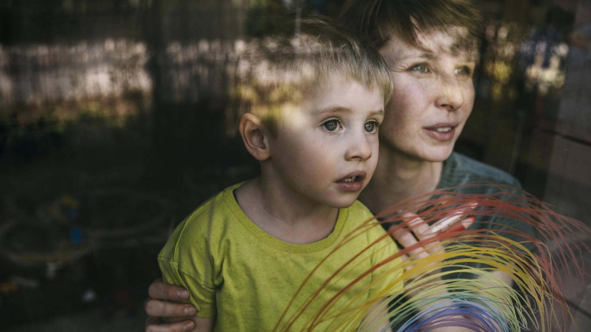 Eine Frau hält einen kleinen Jungen von hinten an den Schultern – beiden schauen zum Fenster hinaus, an das mit dünnen Strichen ein bunter regenbogen gemalt ist. Eine Frau hält einen kleinen Jungen von hinten an den Schultern – beiden schauen zum Fenster hinaus, an das mit dünnen Strichen ein bunter regenbogen gemalt ist.