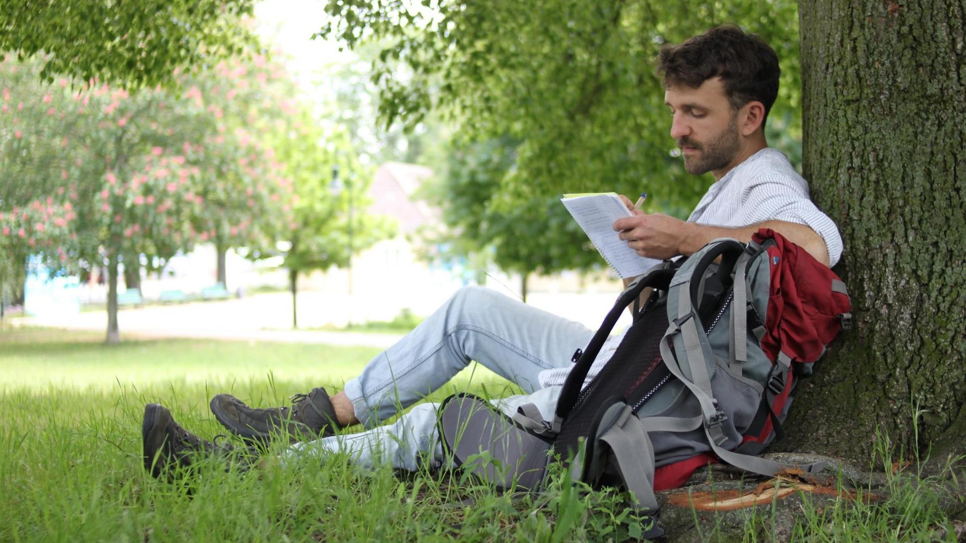 Paul Hildebrandt sitzt an einen Baum gelehnt, auf einen Rucksack gestützt mit einem Notziblock in der Hand auf einer Wiese.