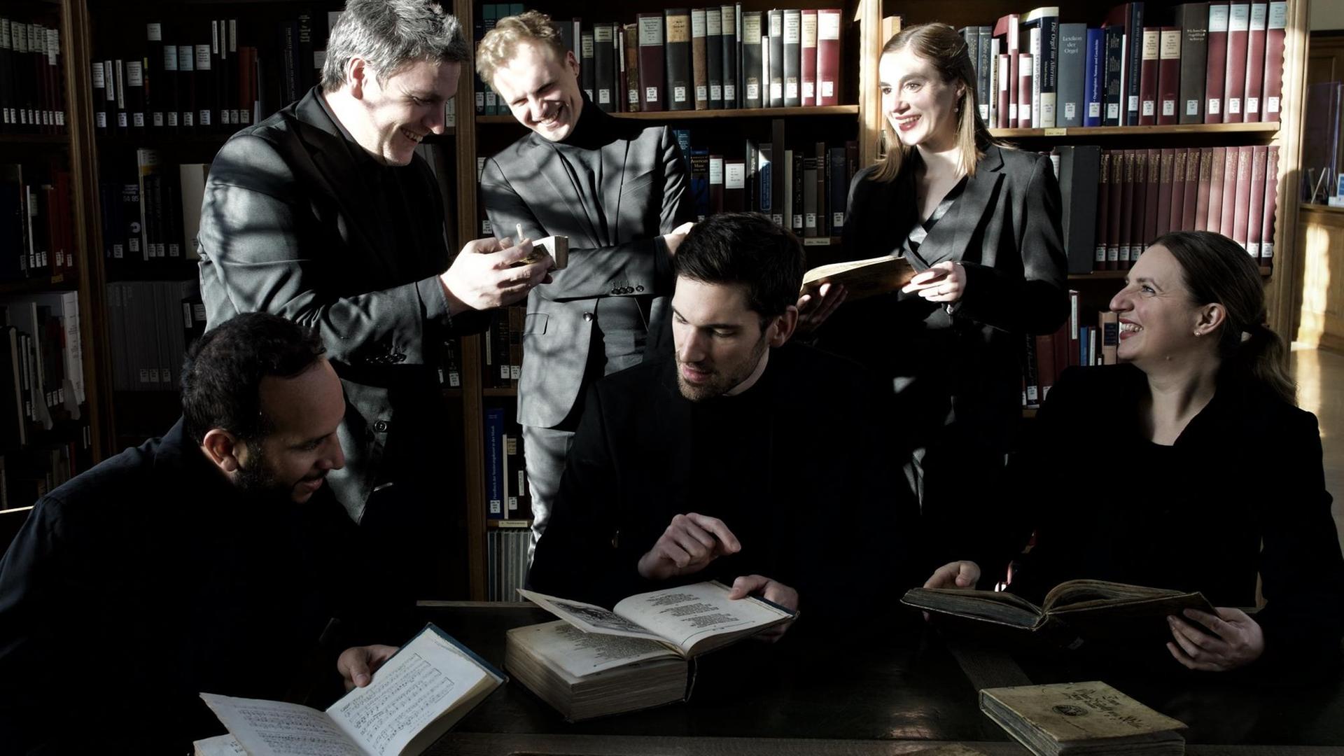 Zwei Frauen und vier Männer stehen und sitzen in einem Raum mit vielen Büchern in Regalen. Sie scheinen sich angeregt zu unterhalten, die meisten blättern in Büchern, alle tragen schwarze Konzertkleidung.