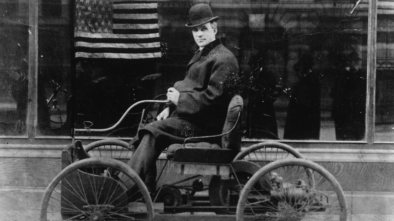 Ein Schwarz-Weiß-Foto zeigt Henry Ford, wie er sein erstes Auto fährt. Im Hintergrund: eine amerikanische Flagge. Henr