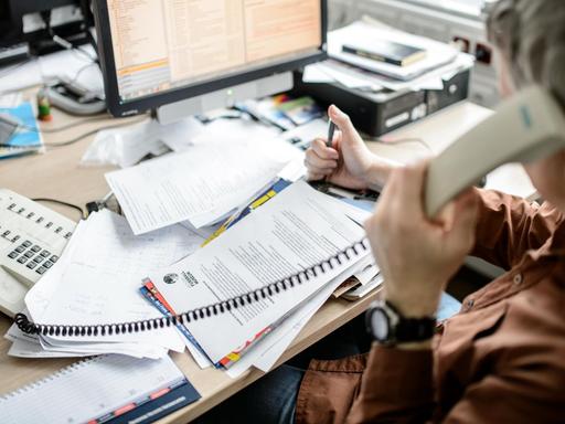 Ein Angestellter sitzt in einem Büro an seinem Schreibtisch und telefoniert vor einem großen Stapel von Unterlagen, aufgenommen am 04.02.2015 in Dresden (Sachsen).