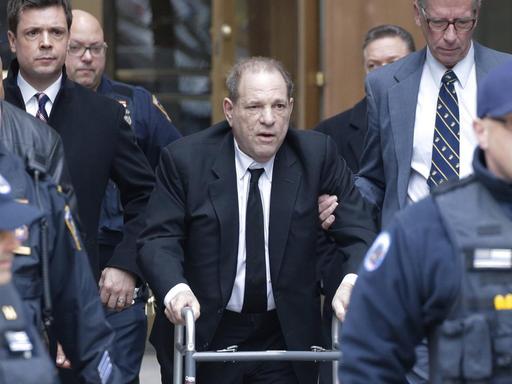 Der Filmproduzent Harvey Weinstein mit Rollator vor einem Gerichtsgebäude in Manhattan. Er wird von einer Person gestützt. Um ihn herum: Polizisten und Anwälte.