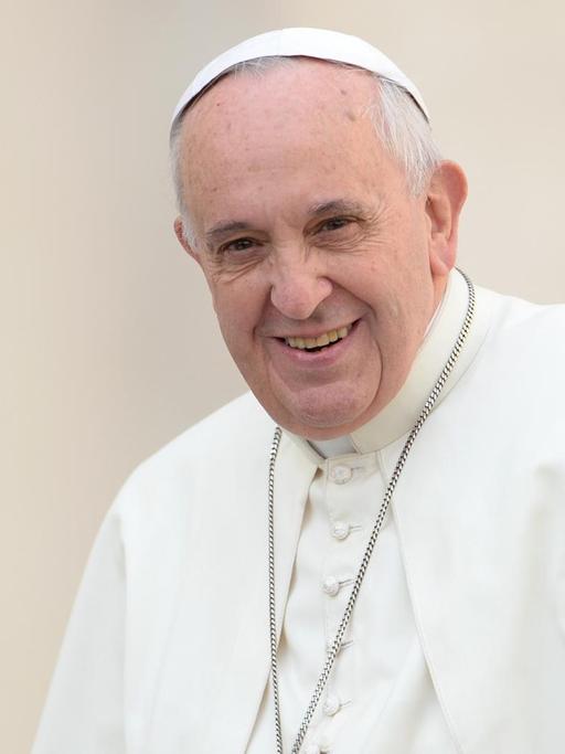 Der Papst lächelt im weißen Gewand und mit umgehängter Kreuzkette vor einer weißen Wand in die Kamera.