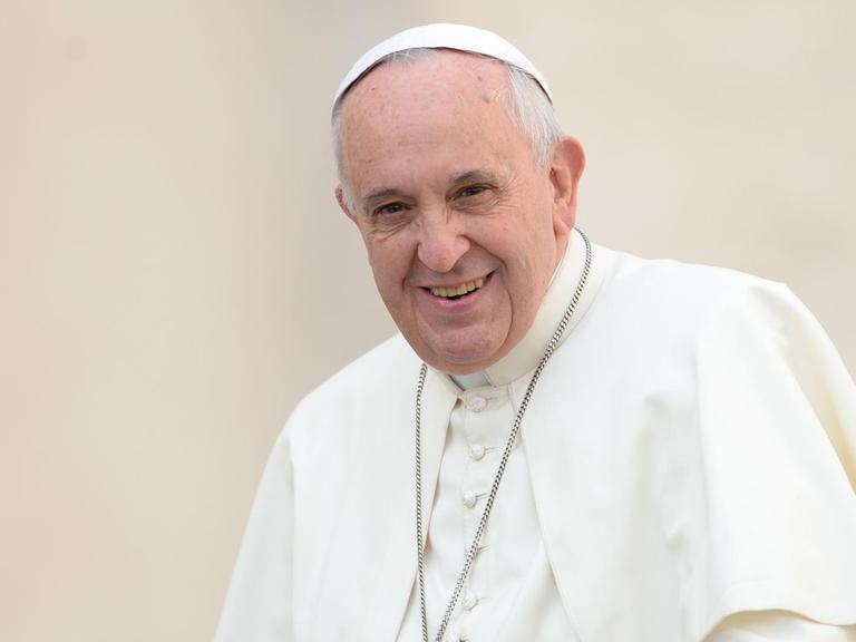 Der Papst lächelt im weißen Gewand und mit umgehängter Kreuzkette vor einer weißen Wand in die Kamera.