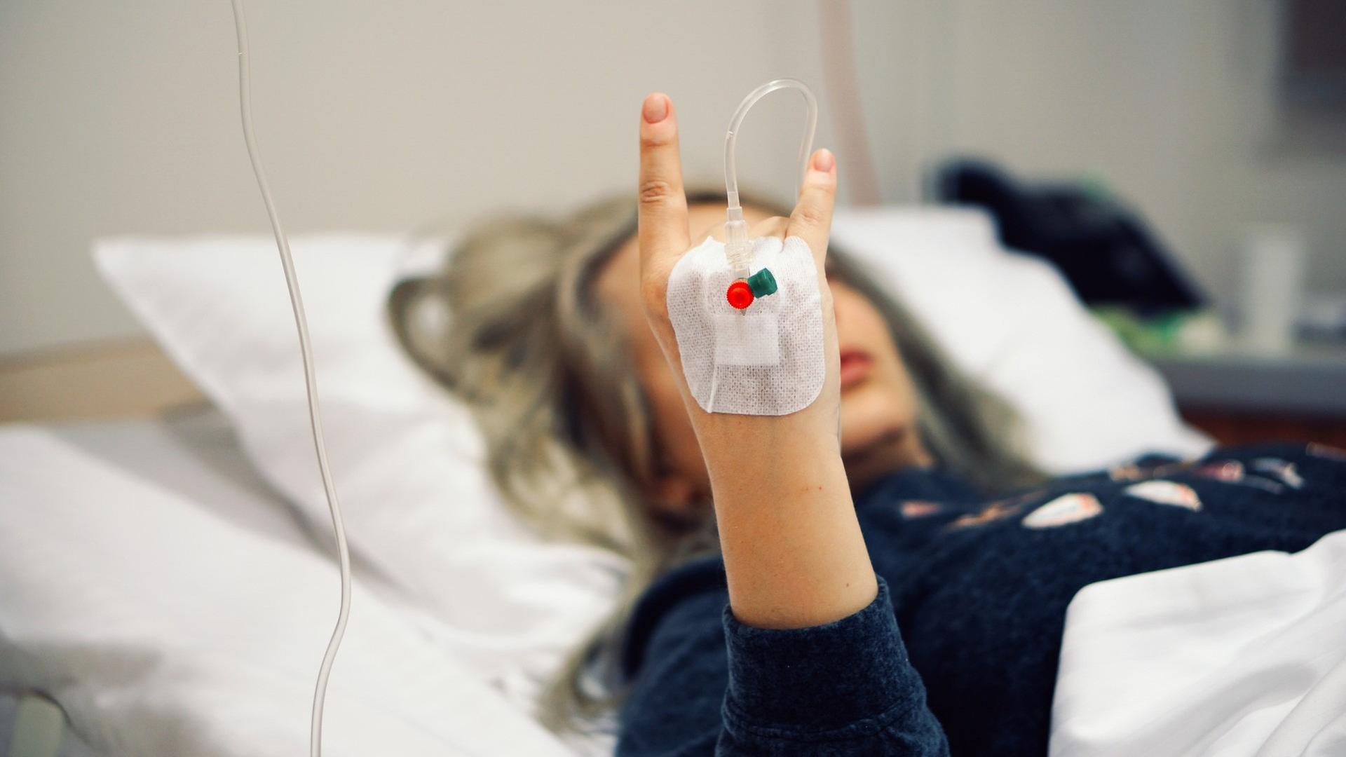 Eine Frau mit einem Tropf liegt im Krankenhausbett und zeigt im Bildvordergrund die mano cornuta. Eine Frau mit einem Tropf liegt im Krankenhausbett und zeigt im Bildvordergrund die mano cornuta.