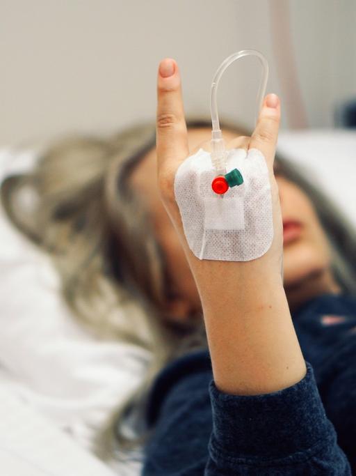 Eine Frau mit einem Tropf liegt im Krankenhausbett und zeigt im Bildvordergrund die mano cornuta.