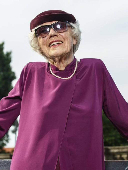 Eine modisch in Violett gekleidete Seniorin mit Perlenkette, Sonnenbrille und Hut posiert vor dem Berliner Fernsehturm