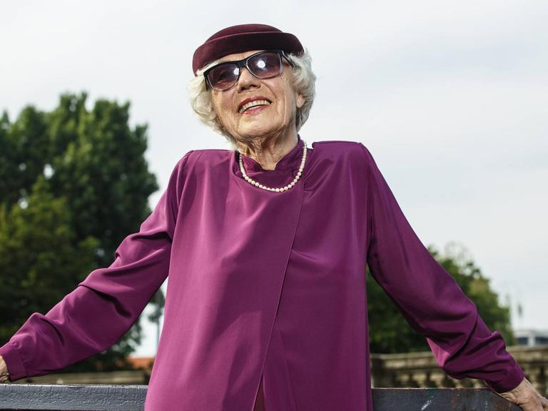 Eine modisch in Violett gekleidete Seniorin mit Perlenkette, Sonnenbrille und Hut posiert vor dem Berliner Fernsehturm
