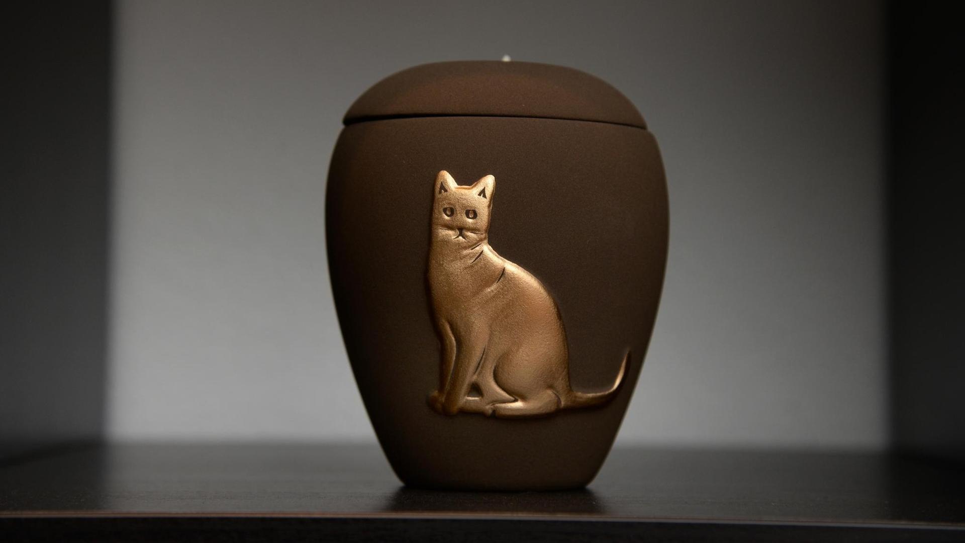 Eine Urne mit dem Relief einer Katze steht in einem Regal. Eine Urne mit dem Relief einer Katze steht in einem Regal.