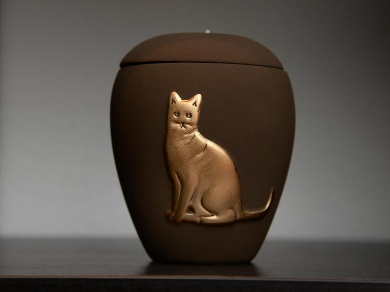 Eine Urne mit dem Relief einer Katze steht in einem Regal.