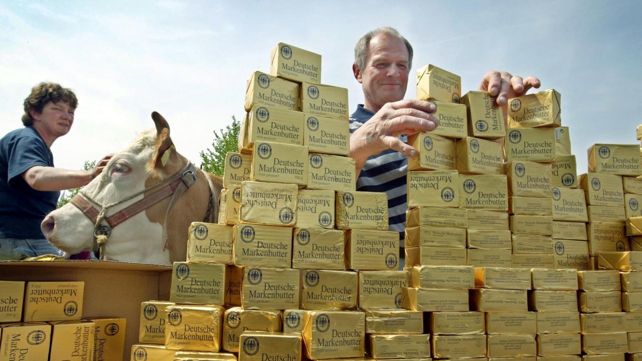 Ein Bauer legt ein Stück Butter auf einen gestapelten Butterberg, im Hintergrund hält eine Bäuerin eine Kuh, aufgenommen 2004 bei Würzburg.