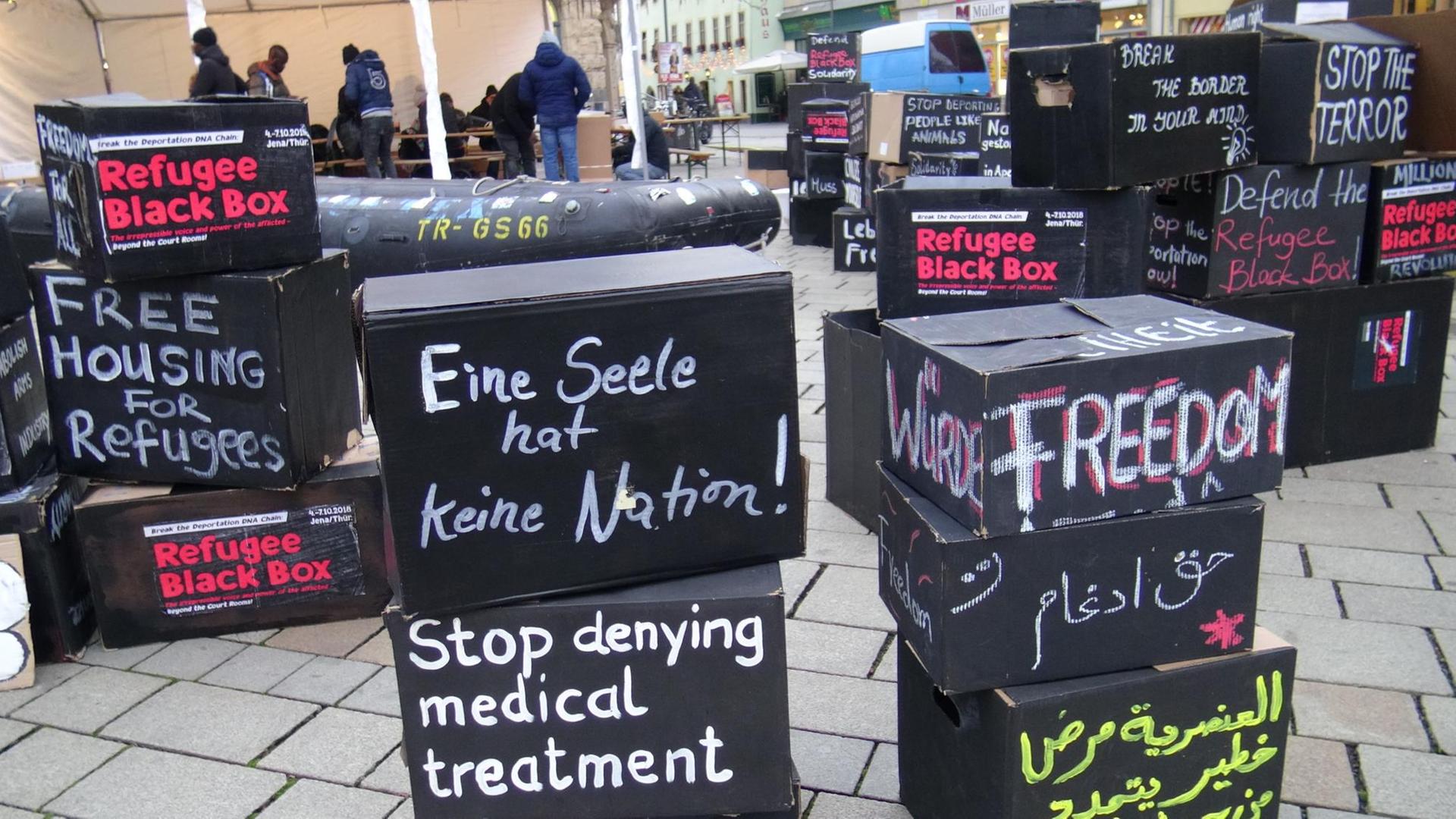 Aufgestapelte schwarze Boxen mit Aufschriften (Refugee Black Box, Eine Seele hat keine Nation!, Stop denying medical treatment, Freedom u.a.), im Hintergrund ein Schlauchboot und mehrere Personen. Aufgestapelte schwarze Boxen mit Aufschriften (Refugee Black Box, Eine Seele hat keine Nation!, Stop denying medical treatment, Freedom u.a.), im Hintergrund ein Schlauchboot und mehrere Personen.