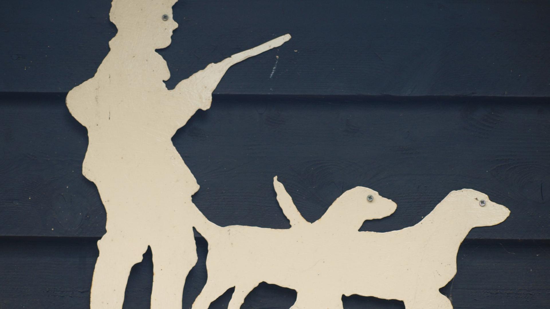 Auf einem Holzschnitt an einem Haus ist ein Jäger mit einem Gewehr und zwei Hunden zu sehen. Auf einem Holzschnitt an einem Haus ist ein Jäger mit einem Gewehr und zwei Hunden zu sehen.